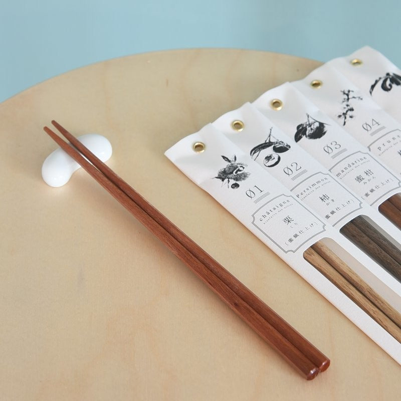 Kawai Tetoca Chopsticks - Peach Wood resting on ceramic holder, with other wood types in elegant packaging in the background.
