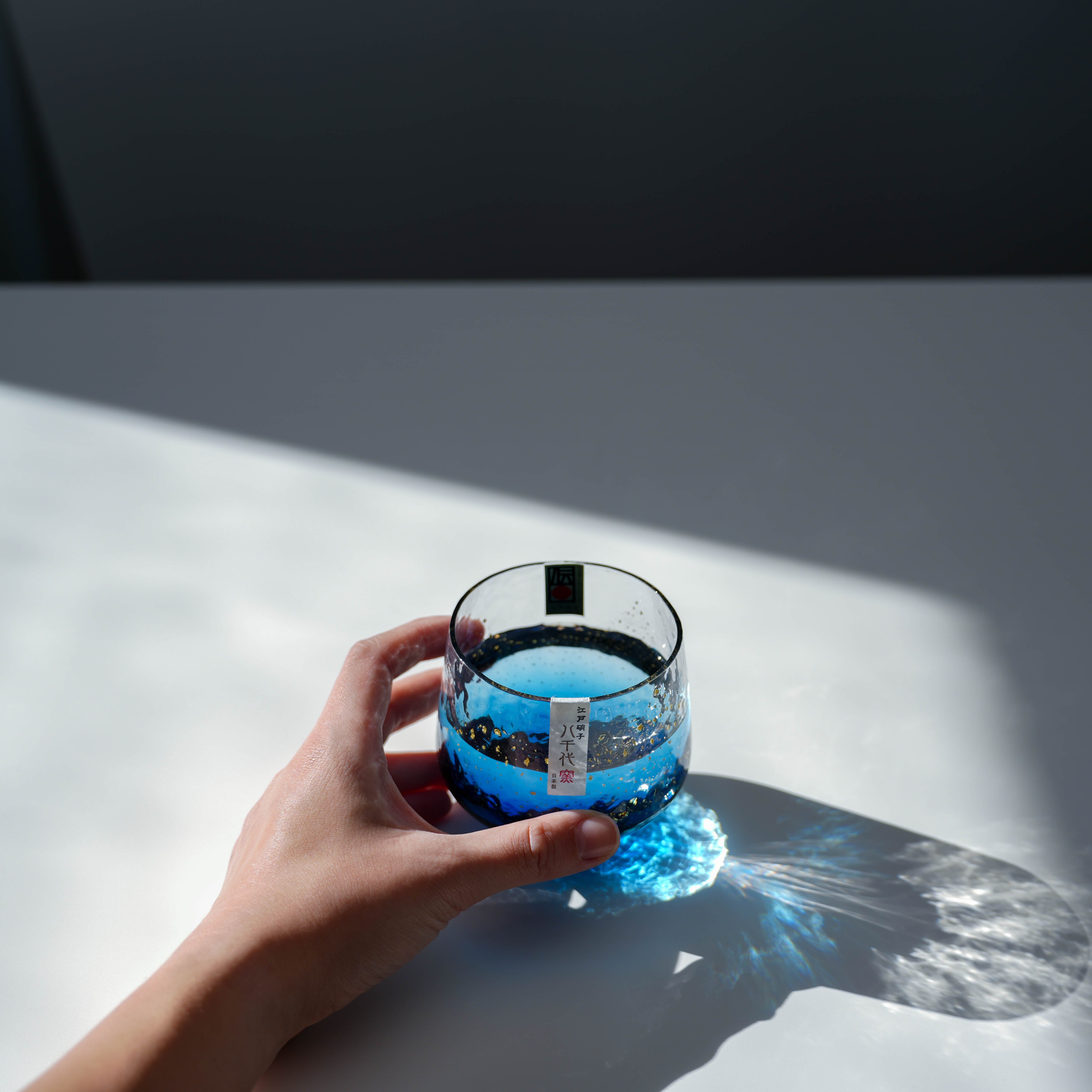 Hand holding Toyo-Sasaki Sapphire Tumbler filled with blue liquid, showcasing Edo glass craftsmanship and light reflection.