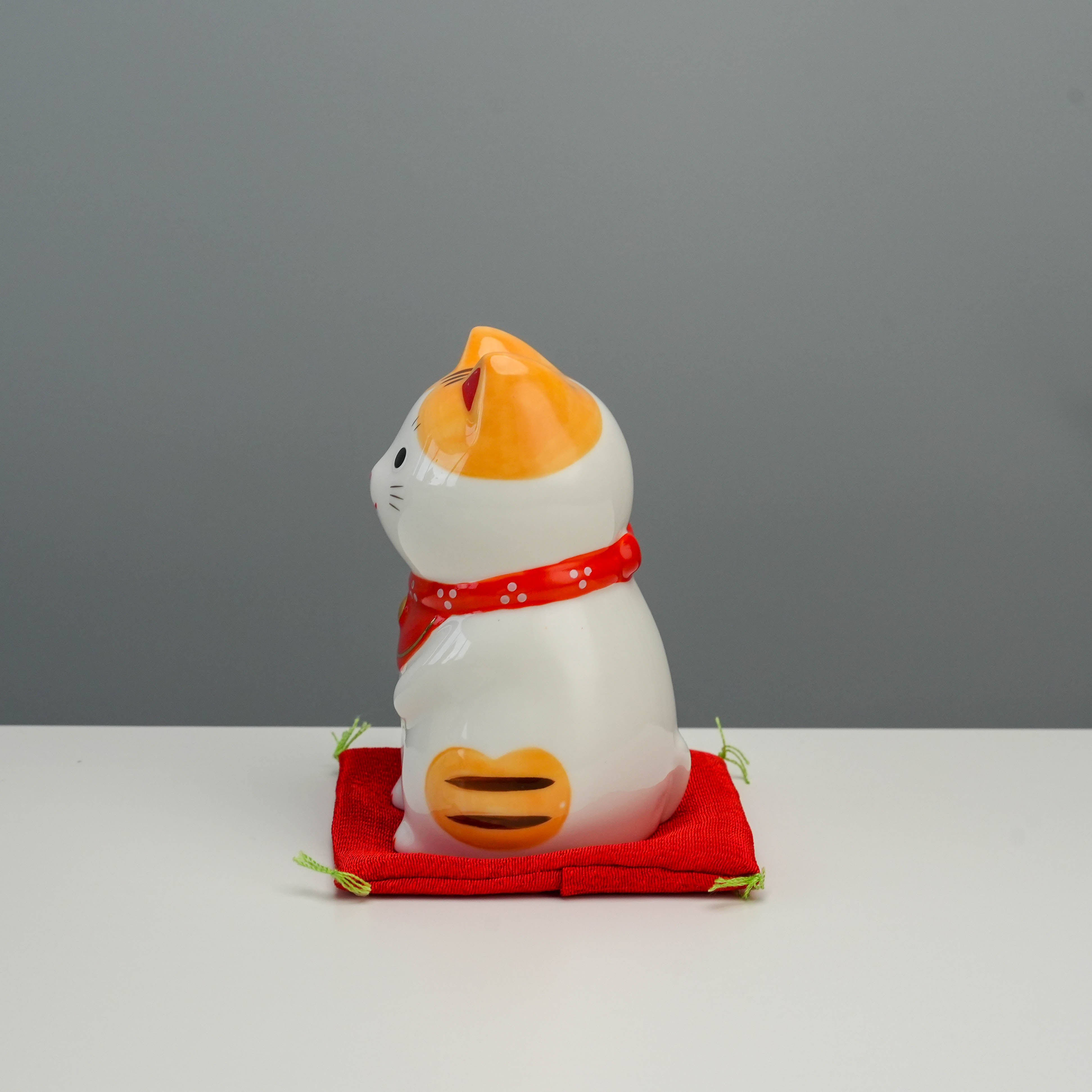 Side view of Yakushigama Lucky Cat - Japanese Fortune Charm with orange tiger stripes and red scarf, placed on decorative red cushion.