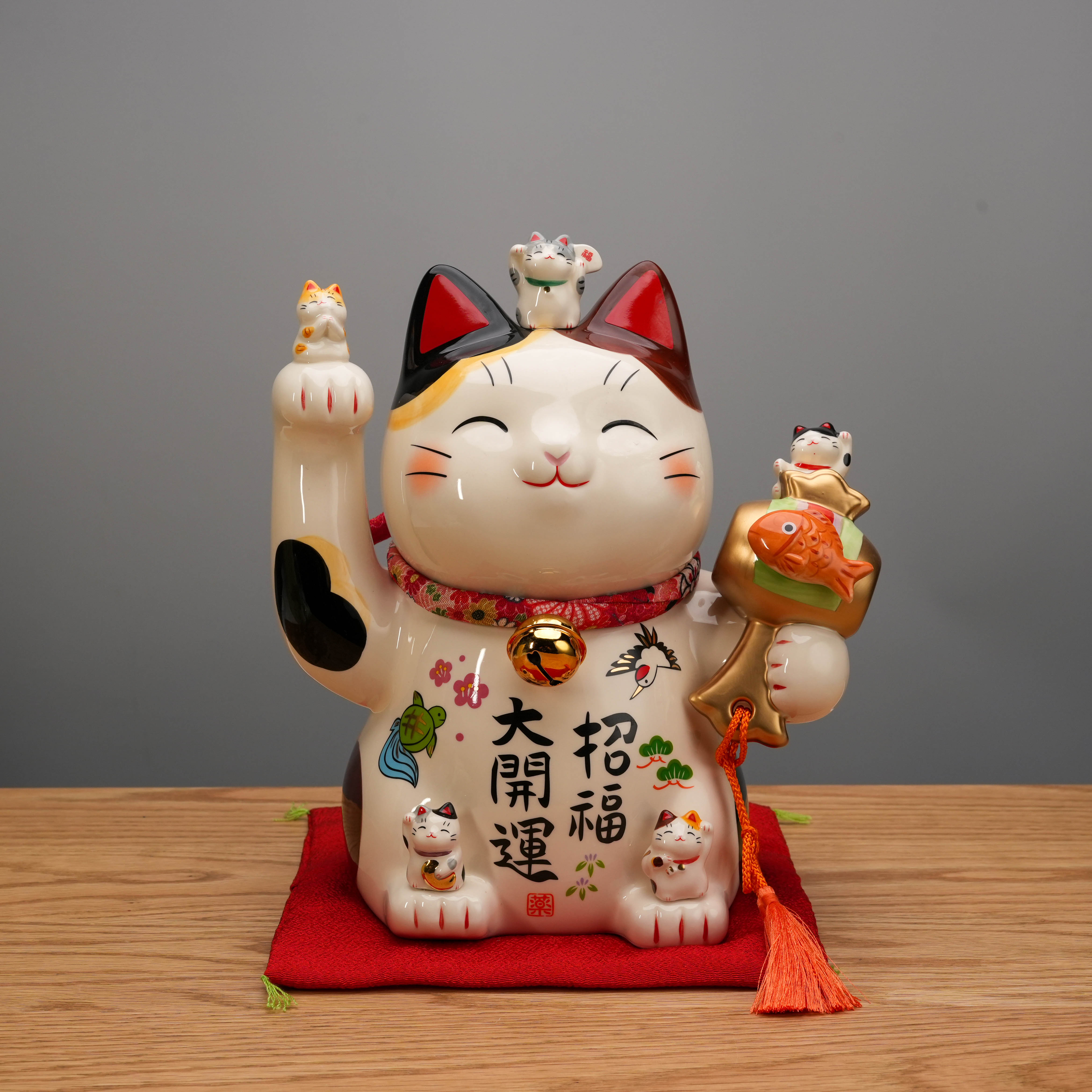 Yakushigama Bliss Beckoning Cat figurine with raised paw, gold bell, and fish accessory on red cushion, symbolizing prosperity and happiness.
