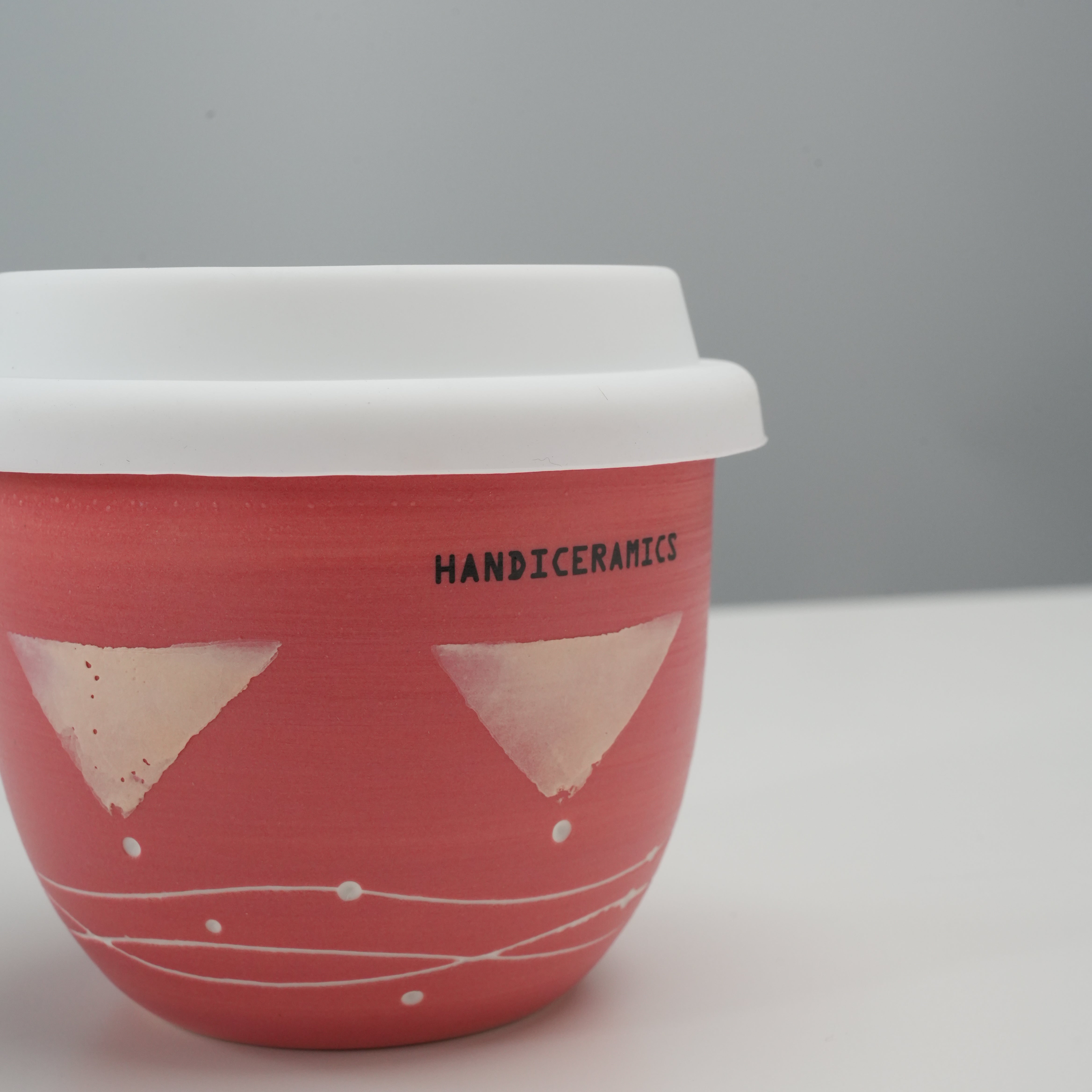 Close-up of Handiceramics Travel Cup - Ceramic Coffee Cup with Lid showing handmade strawberry pink finish and brand logo, perfect for stylish sipping.