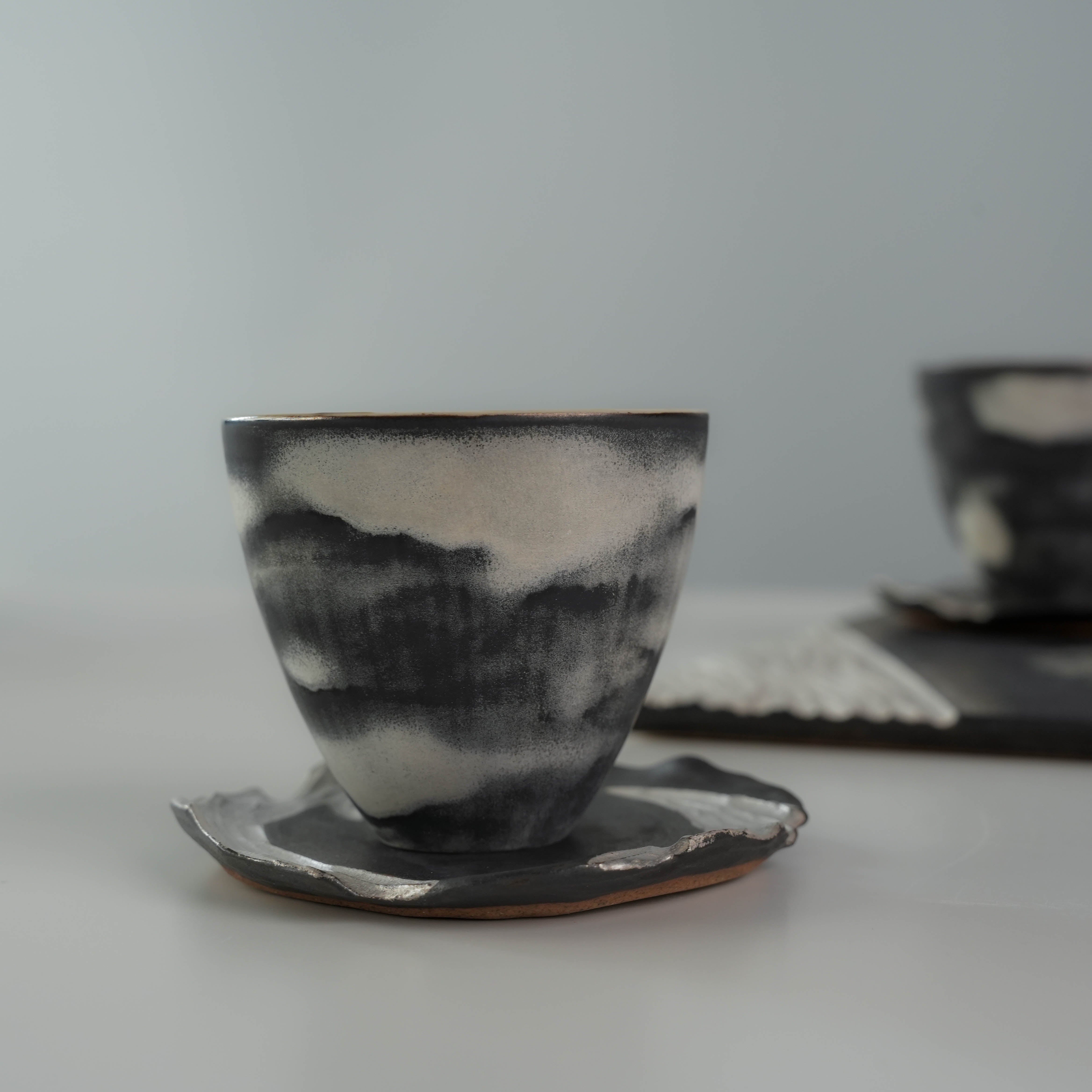 Handcrafted tea cup from Lonesome Studio Gushan Tea Set with cloudy black and white glaze, resting on a silver-edged saucer.