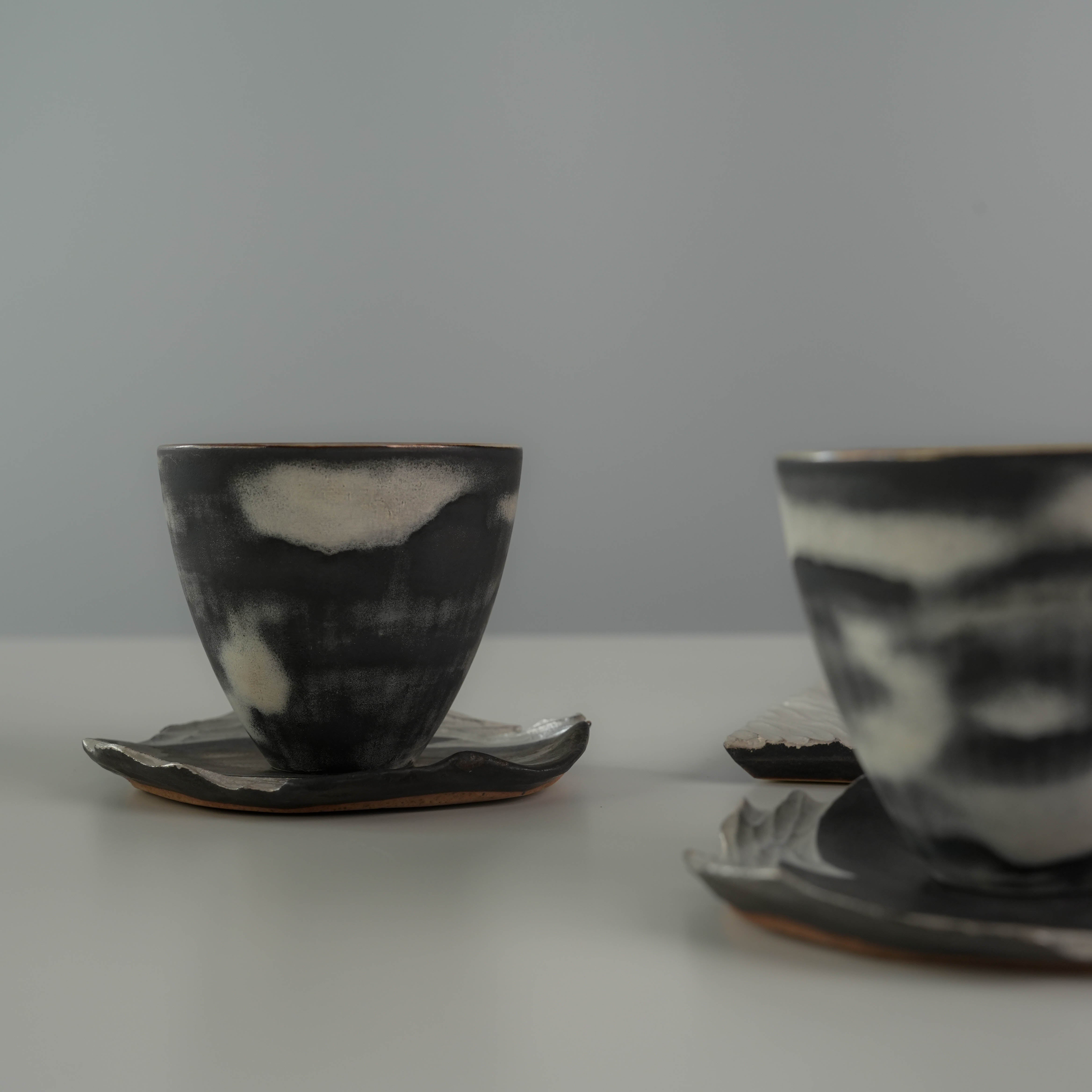 Two handmade tea cups from the Lonesome Studio Gushan Tea Set, featuring unique black and white glaze with silver-accented saucers.
