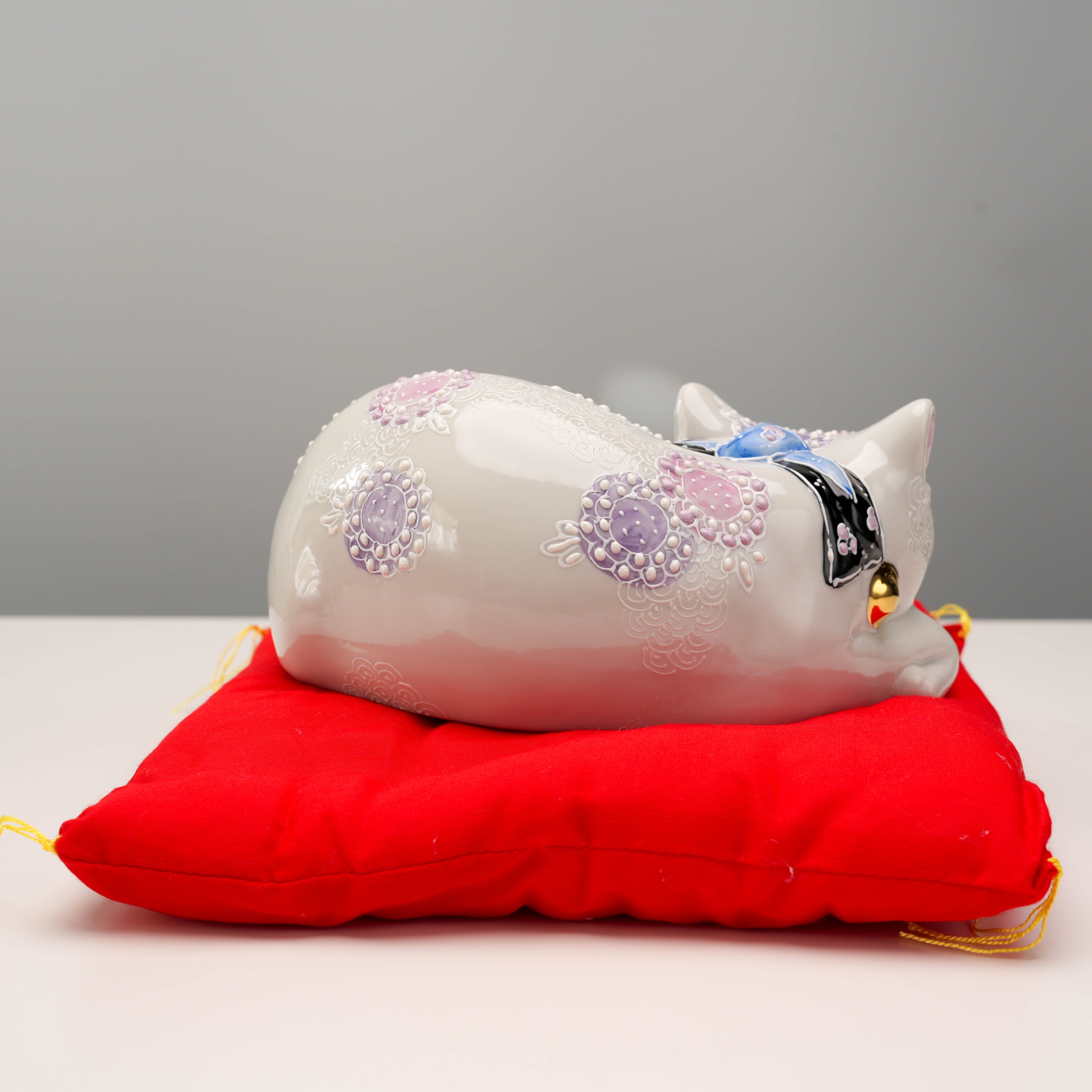 Side view of Kutani Ware Sleeping Cat figurine with floral motifs and golden bell, placed on red cushion for elegant home display.