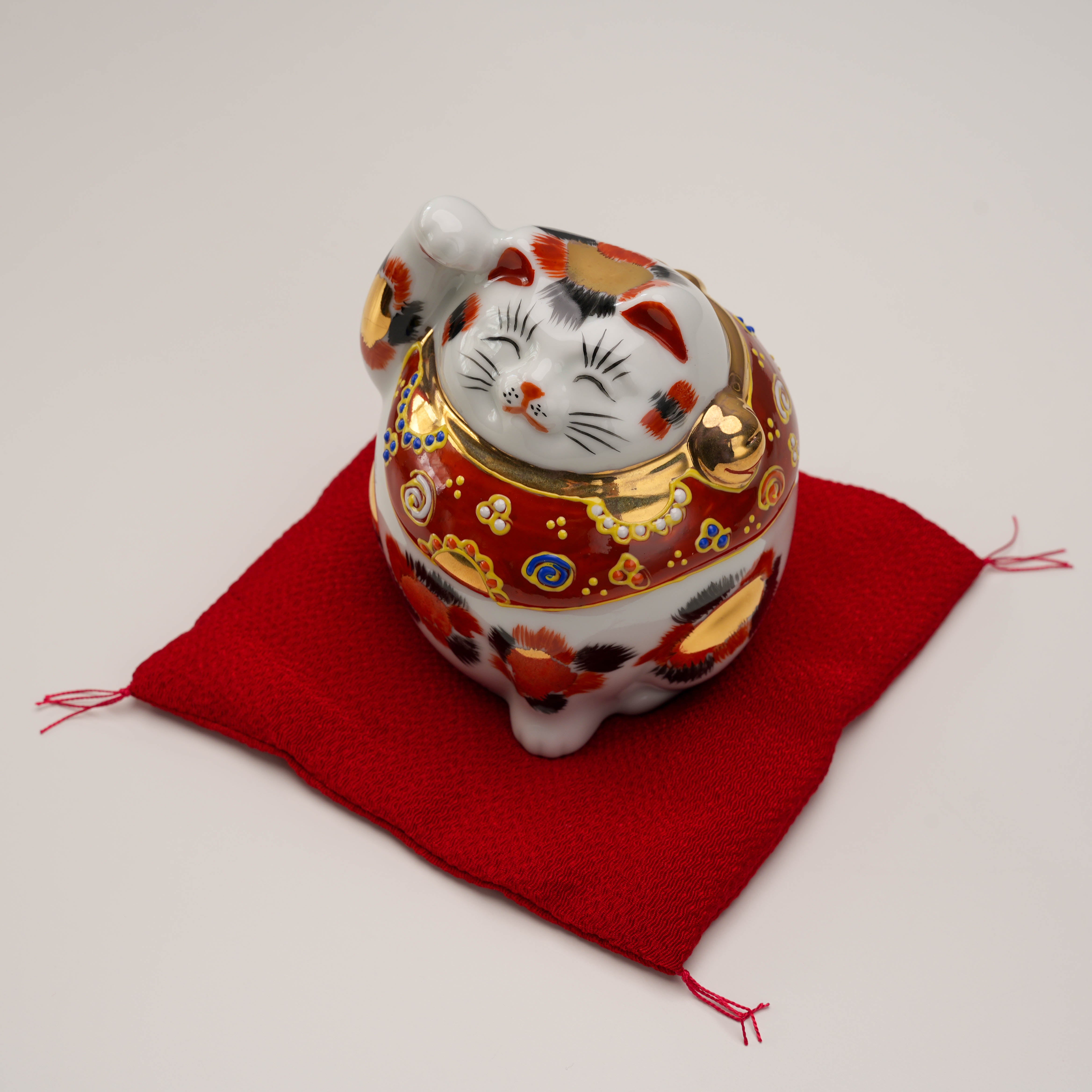 Top view of Kutani Ware Cat – Lucky Calico ornament on red cushion, showcasing its rounded form and detailed collar design.