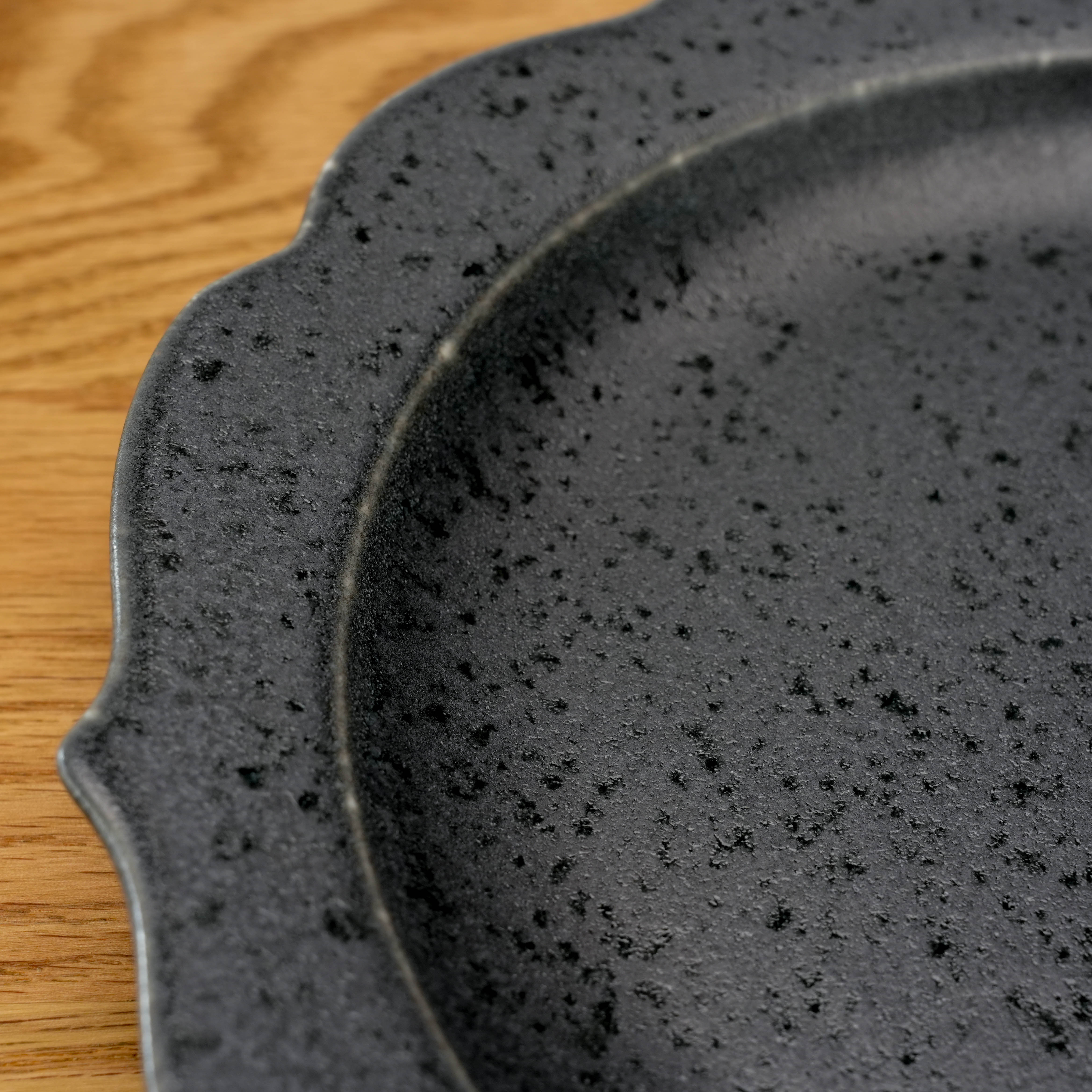 Close-up of Kikyo Black Plate showing speckled matte glaze and scalloped edge. Mino Ware craftsmanship for stylish dinnerware collections.