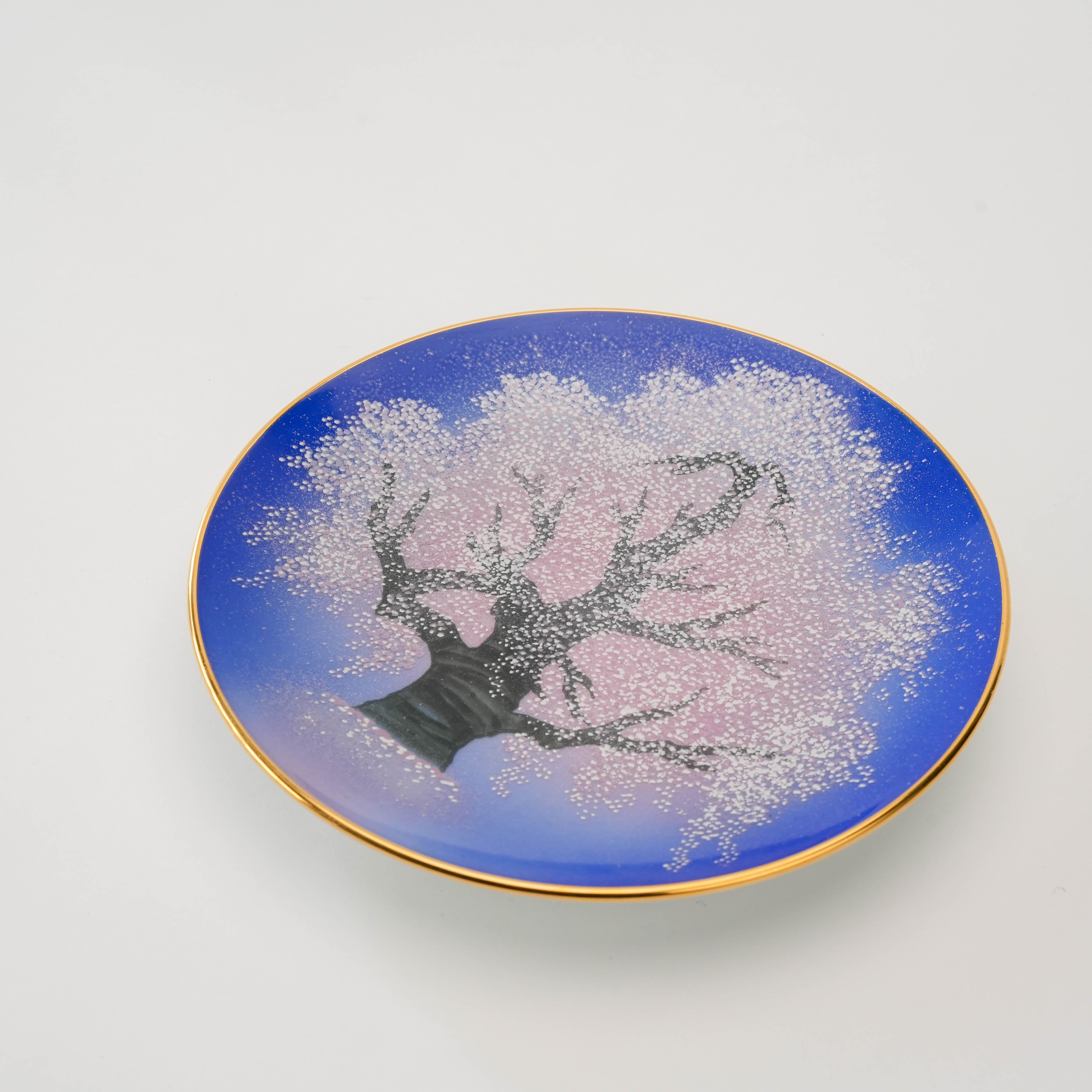 Angled view of the Arita Yozakura Cherry Blossom saucer with hand-painted sakura tree and gold-rimmed edge on a blue gradient background.
