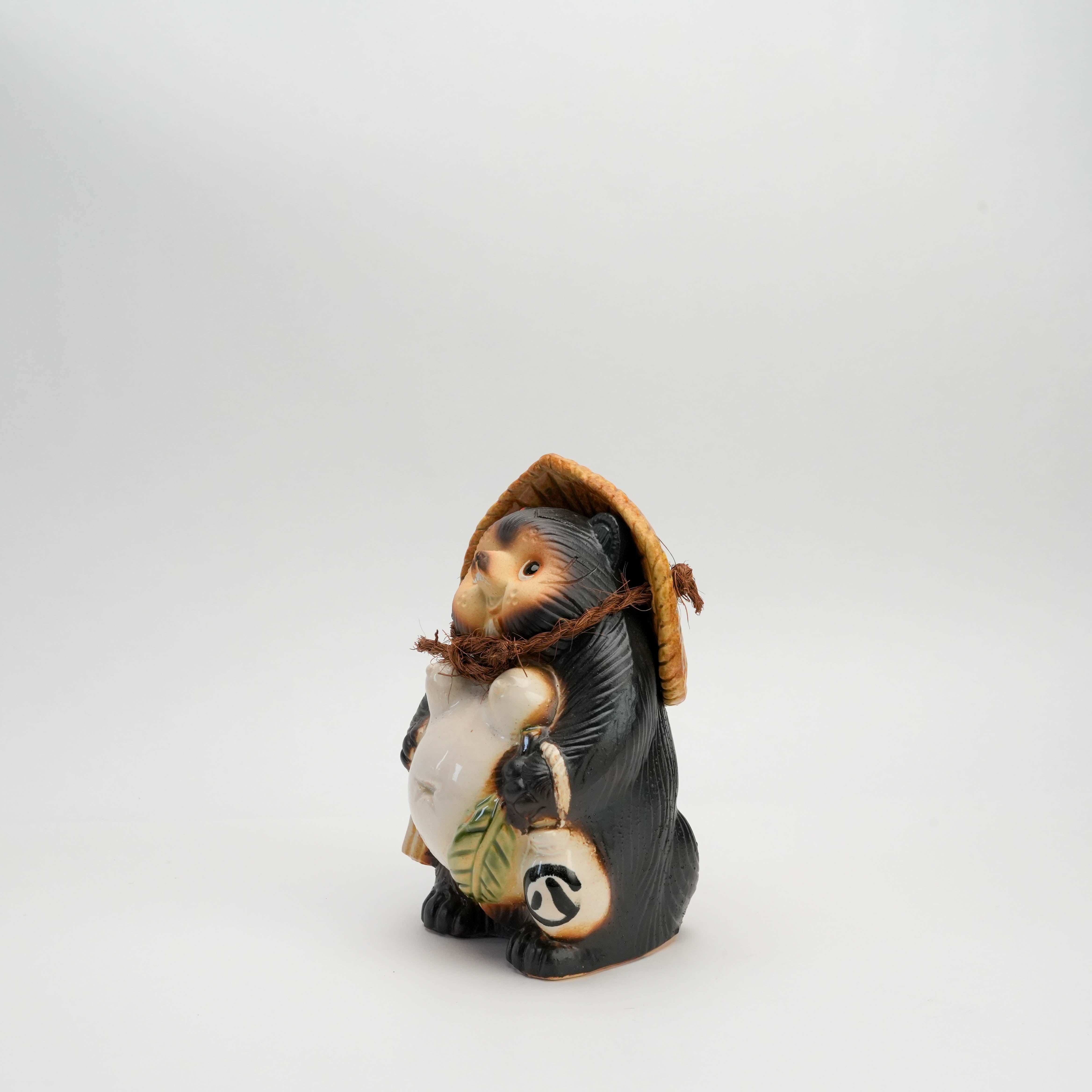 Angled view of handmade Shigaraki Ware Tanuki Ornament holding a sake bottle and wearing a straw hat. Symbol of prosperity and charm.