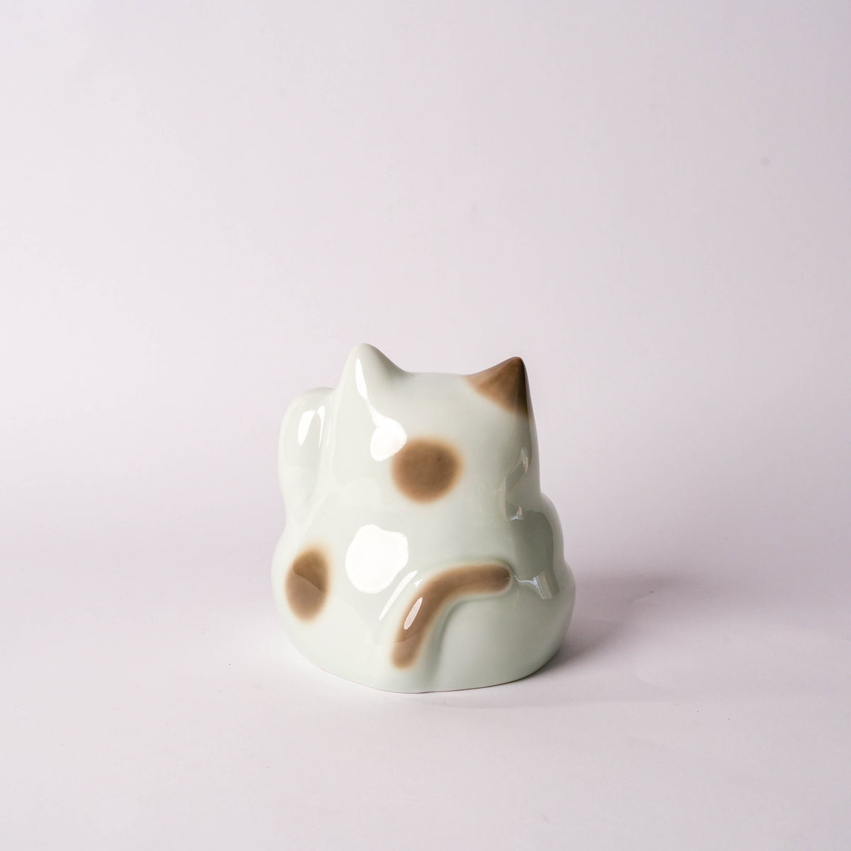 Side view of ceramic cat figurine with brown spots, sitting on a white surface against a plain white background, decorative home decor