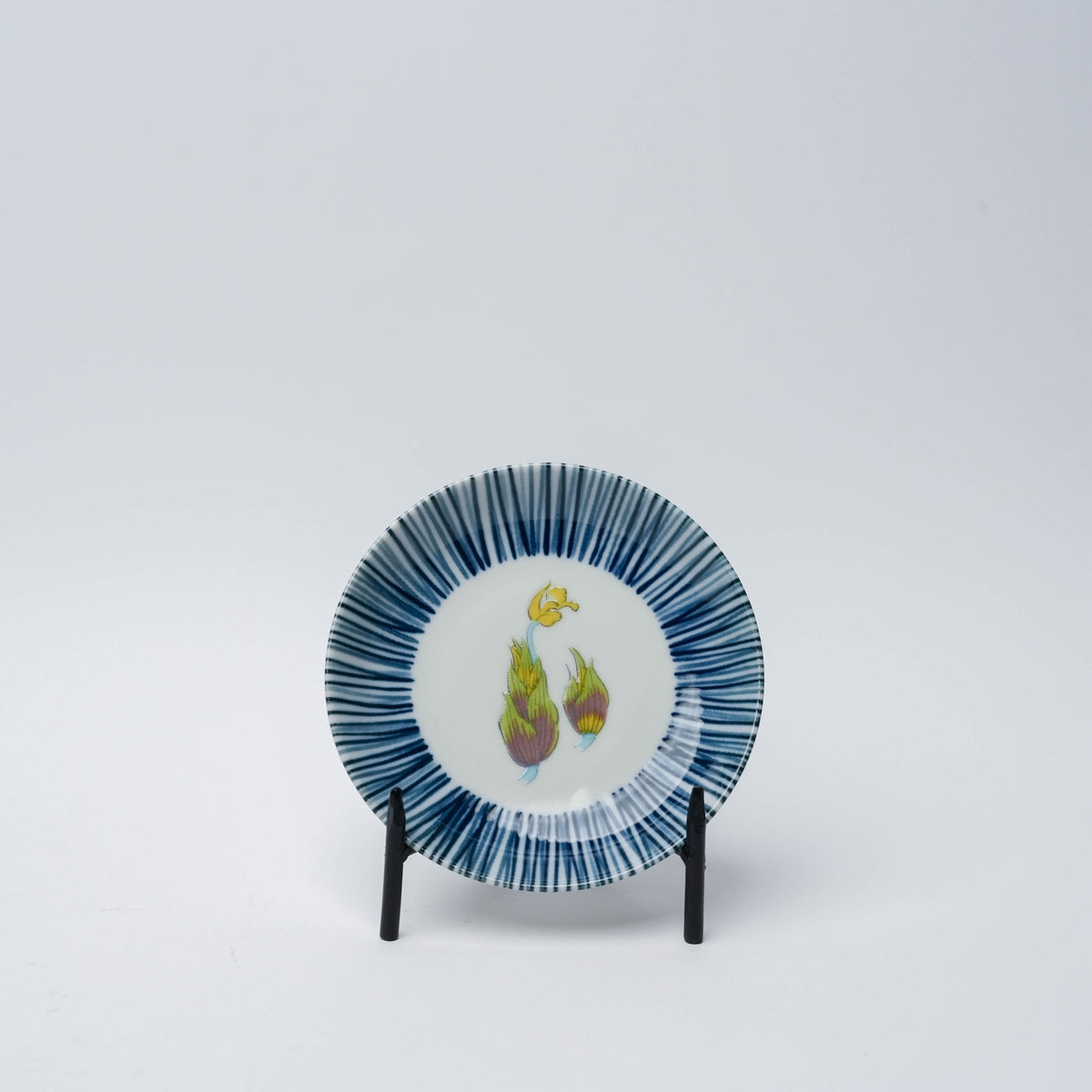 Porcelain small plate with blue striped border and colorful vegetable motif featuring artichokes, set on black display stand against white background.