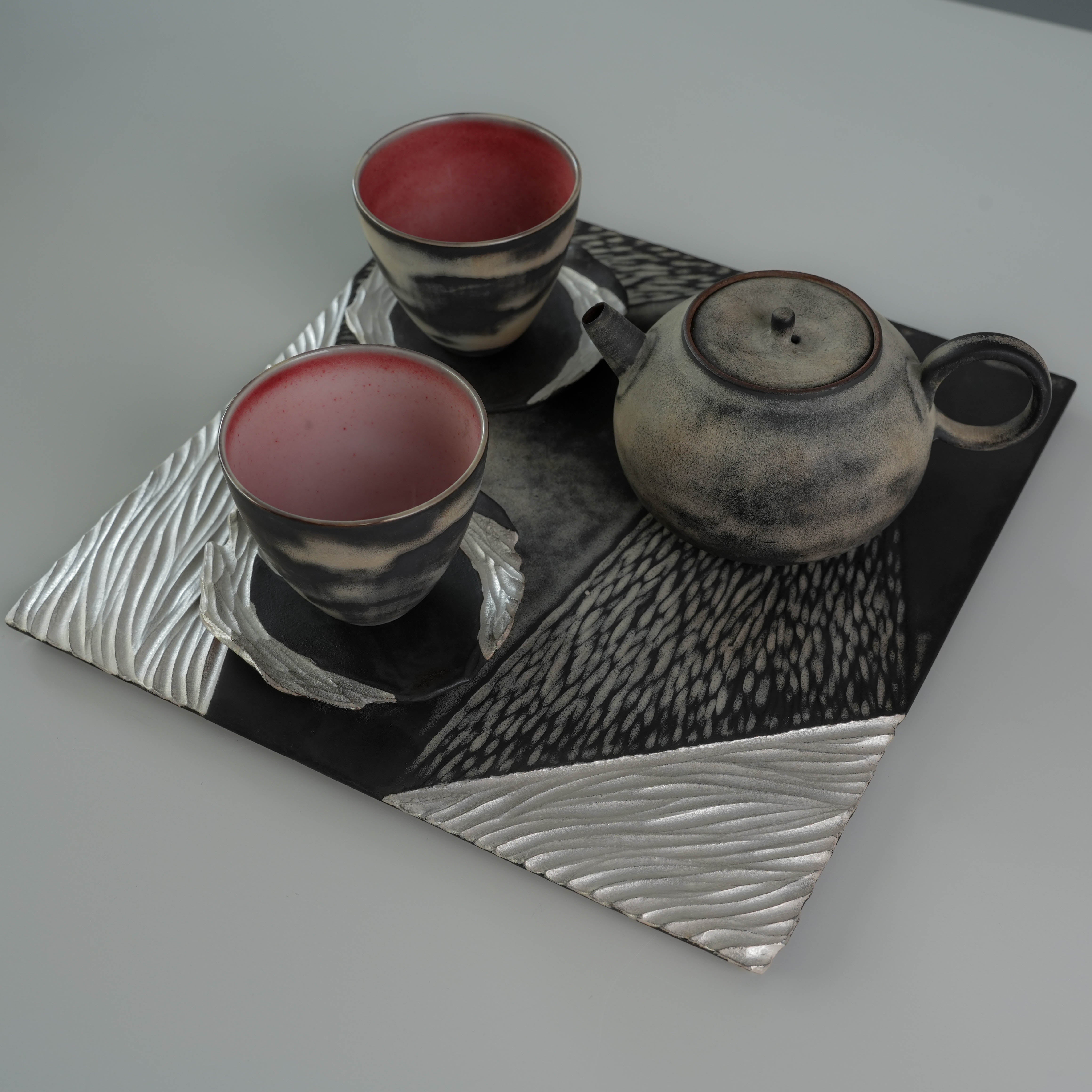 Angled view of Lonesome Studio Gushan Tea Set featuring teapot and two cups on a textured black and silver tray.