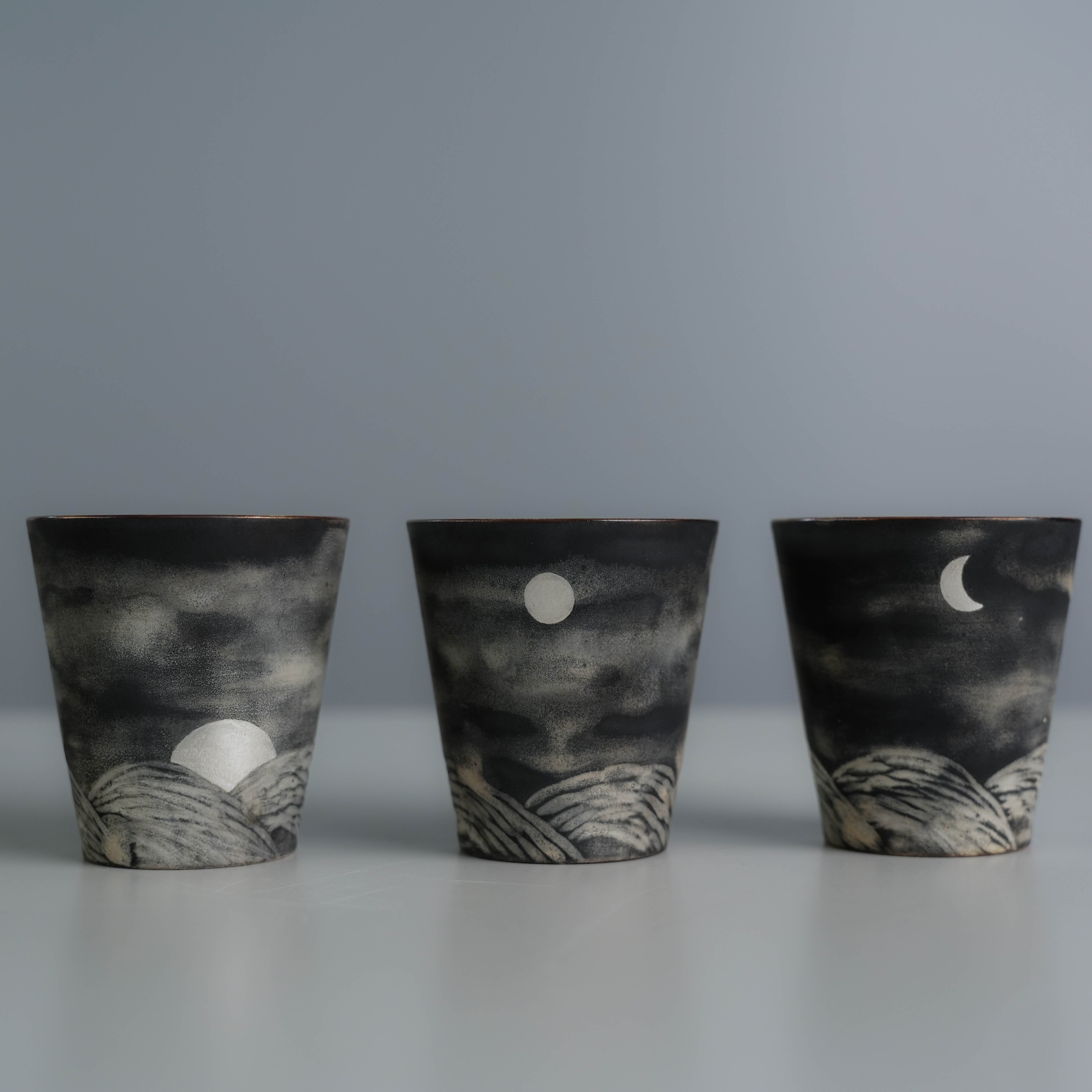 Three Lonesome Studio Qushui tea cups with full moon, crescent, and sunrise motifs, handmade with silver glaze for Gongfu Cha tea ceremonies.