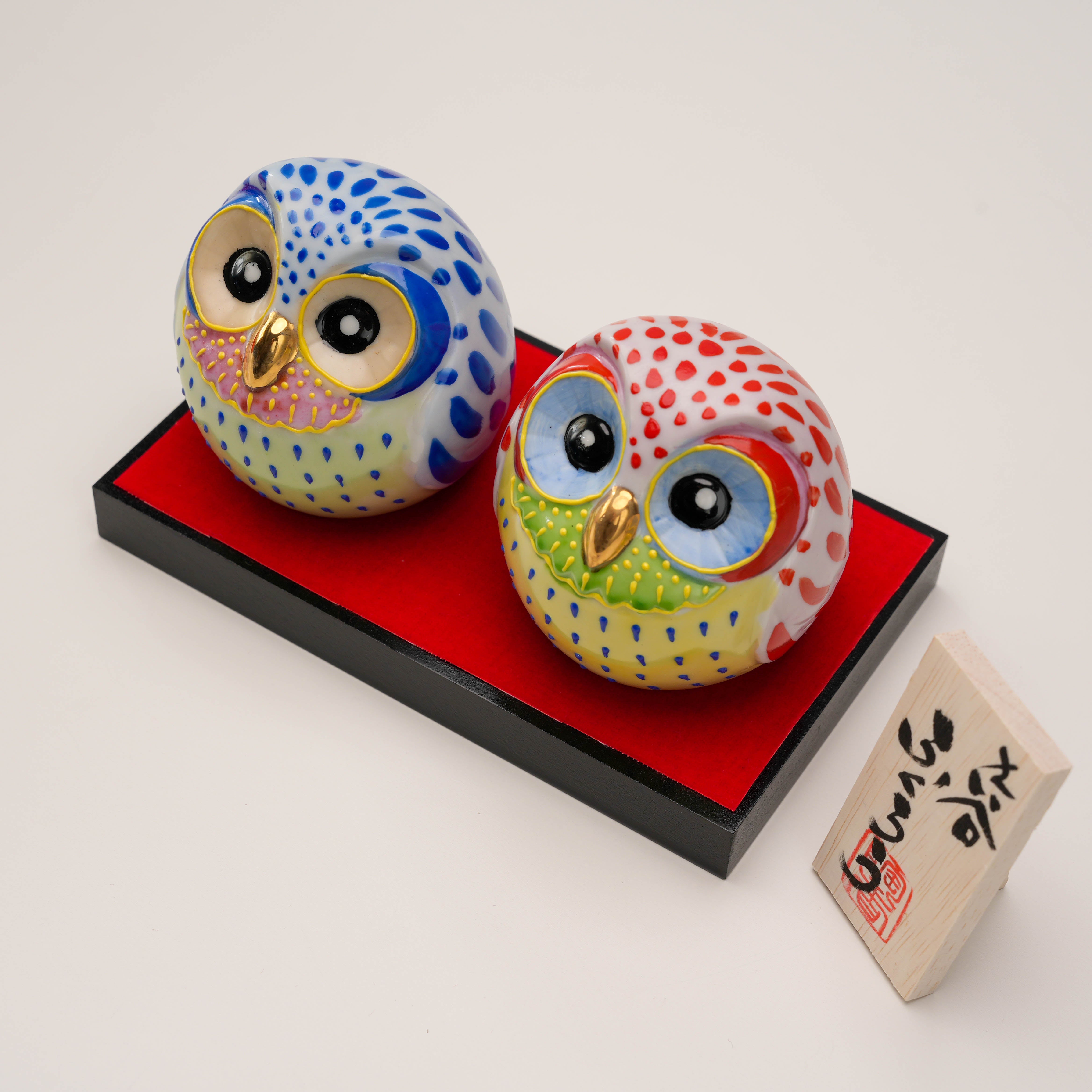 Vibrant Kutani Ware owl figurines in red and blue with glossy finish, displayed on a red velvet base with wooden name plaque. Symbol of good fortune.