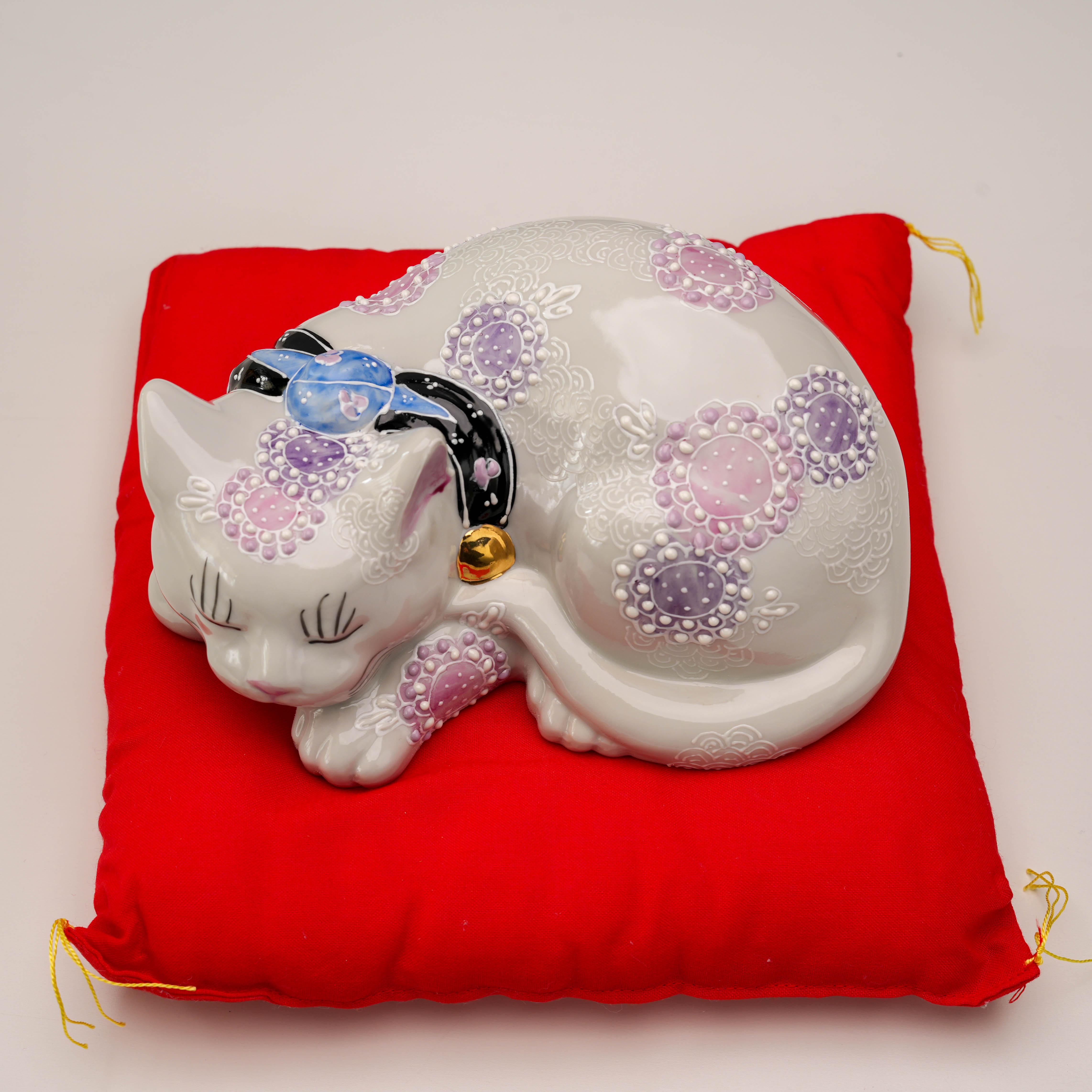 Overhead angle of Kutani Ware Sleeping Cat with delicate purple floral designs, displayed on red cushion for decorative charm.